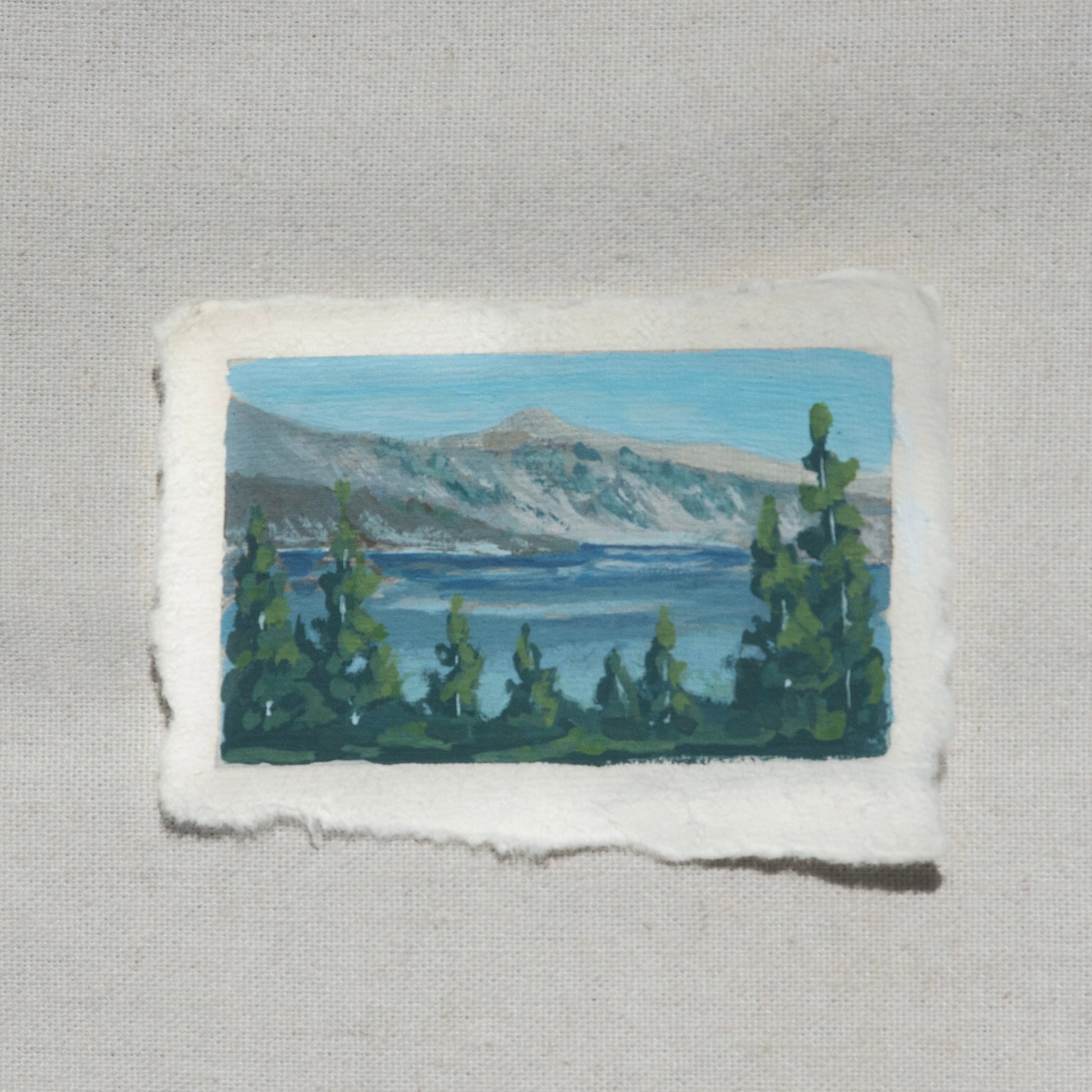 Mini gouache painting of the mountains, pine trees, and vibrant blue water of Crater Lake by Laura Van Moorleghem from Crater Lake National Park