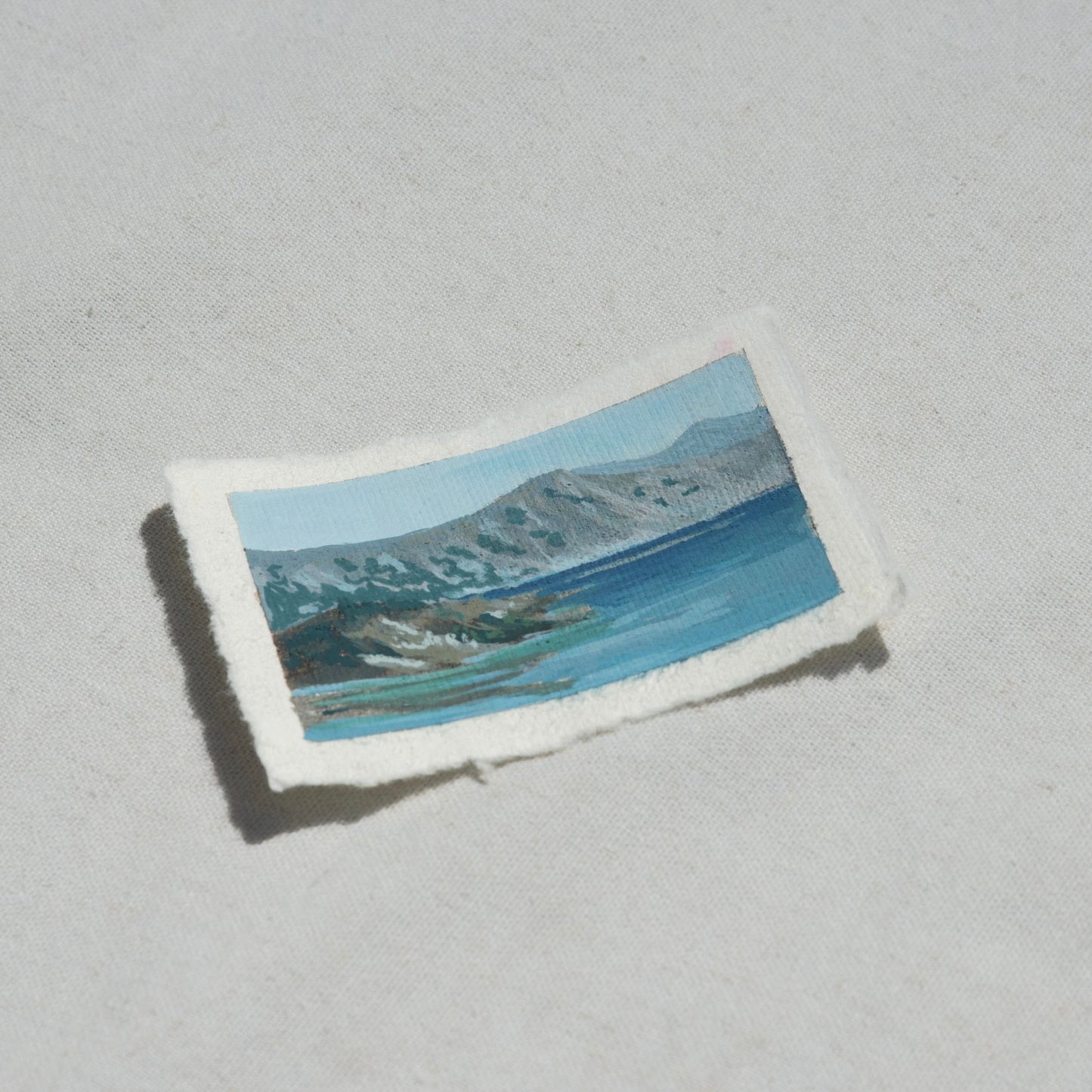 Small rectangular painting of crater lake landscape with mountains and vibrant blue lake water on 100% cotton deckled edge paper