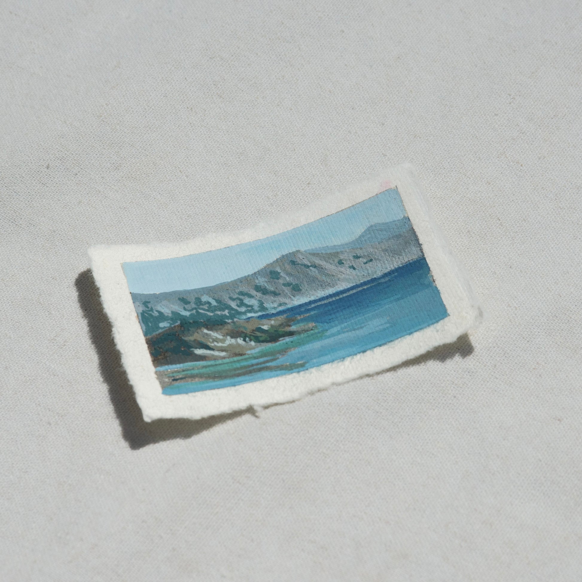 Small rectangular painting of crater lake landscape with mountains and vibrant blue lake water on 100% cotton deckled edge paper